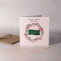 Christmas card on pink background with wreath design, red bows and pink baubles. Personalised calligraphy name.