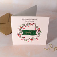 Christmas card with floral wreath and personalised name on a pink background