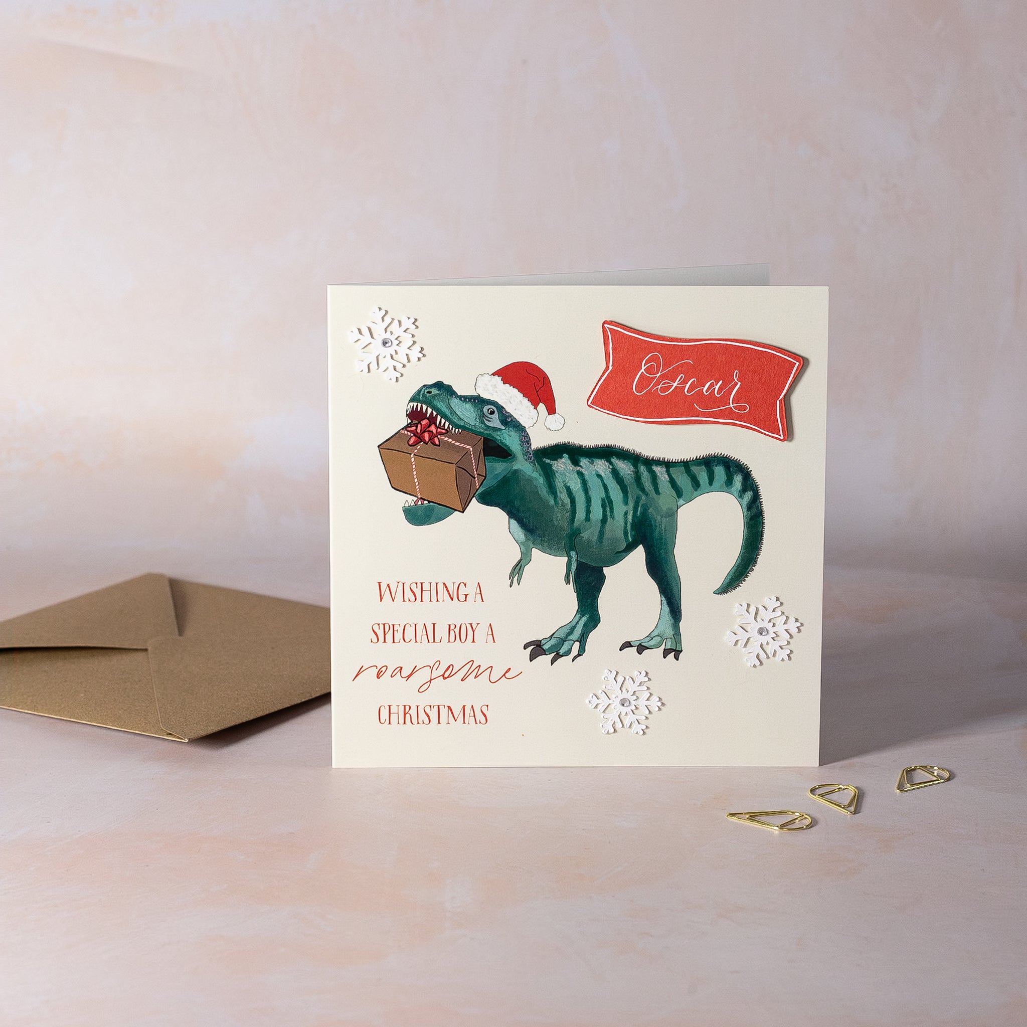 Christmas card with a dinosaur holding a gift, wearing a Santa hat, on a beige background.