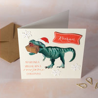 Christmas card with a dinosaur holding a gift and wearing a Santa hat, on a light background.