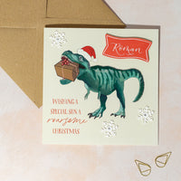 Christmas card with a dinosaur holding a gift and wearing a Santa hat, snowflake decorations and personalised name