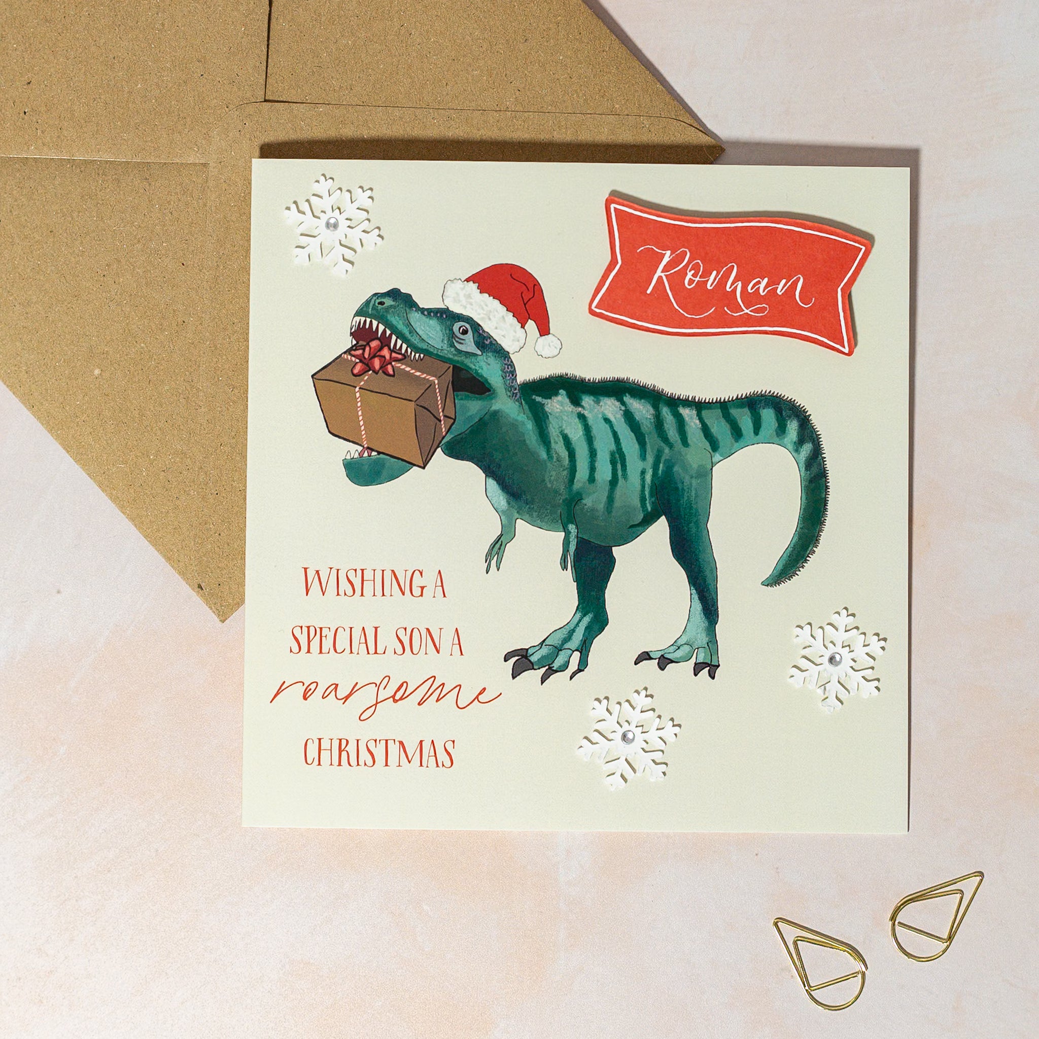 Christmas card with a dinosaur holding a gift and wearing a Santa hat, snowflake decorations and personalised name