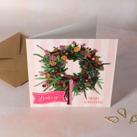 Christmas card with a wreath design and 'Merry Christmas' with personalised name on pink card.